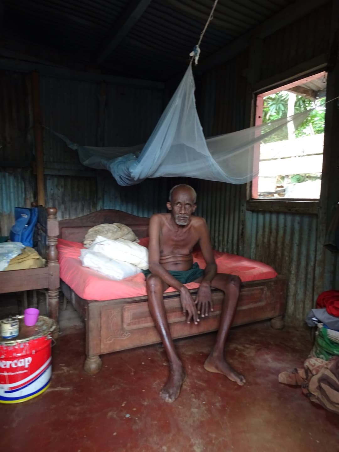 Homme âgé assis sur son lit, sous une moustiquaire, dans son habitation.