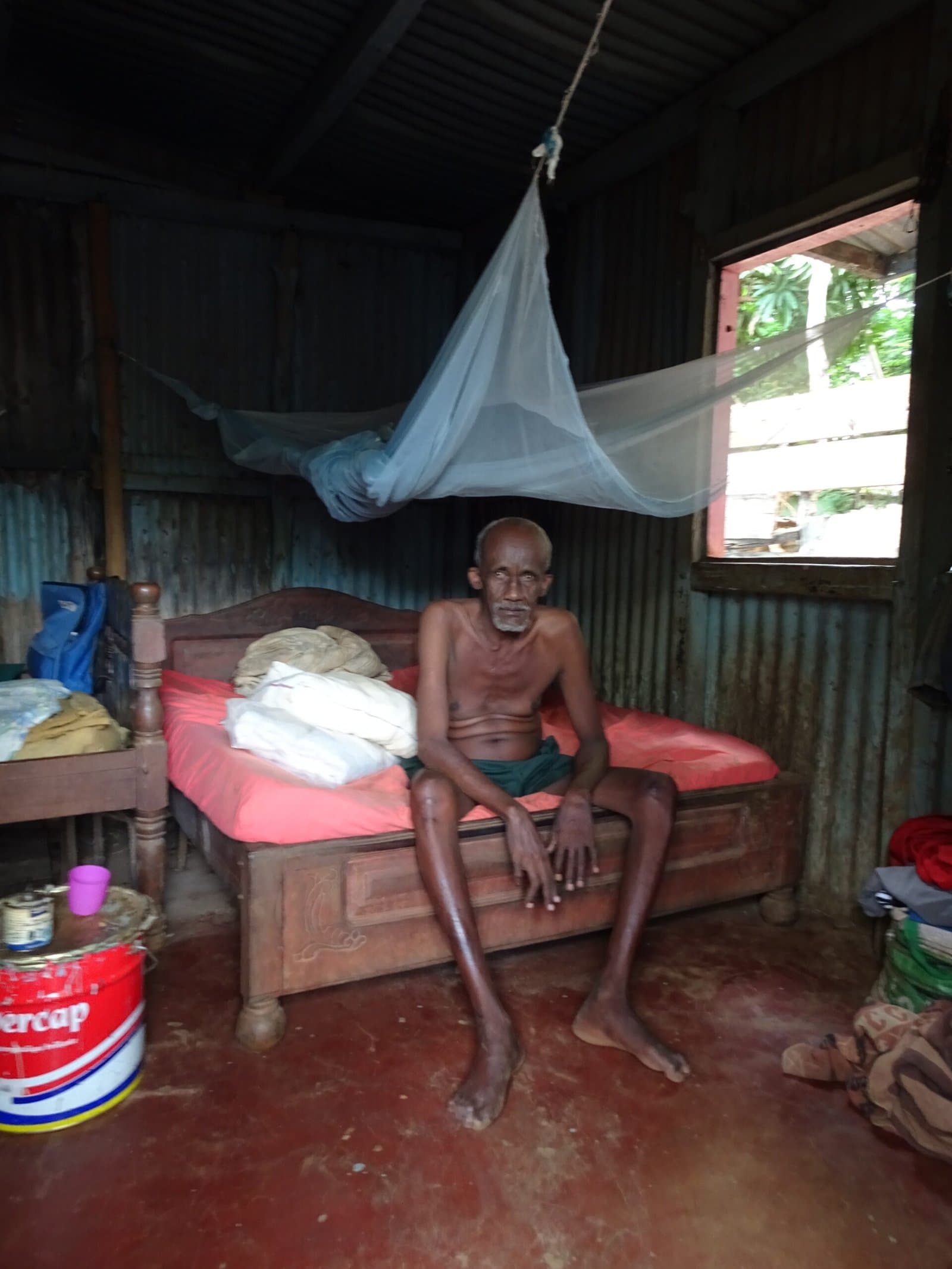 Homme âgé assis sur son lit, sous une moustiquaire, dans son habitation.