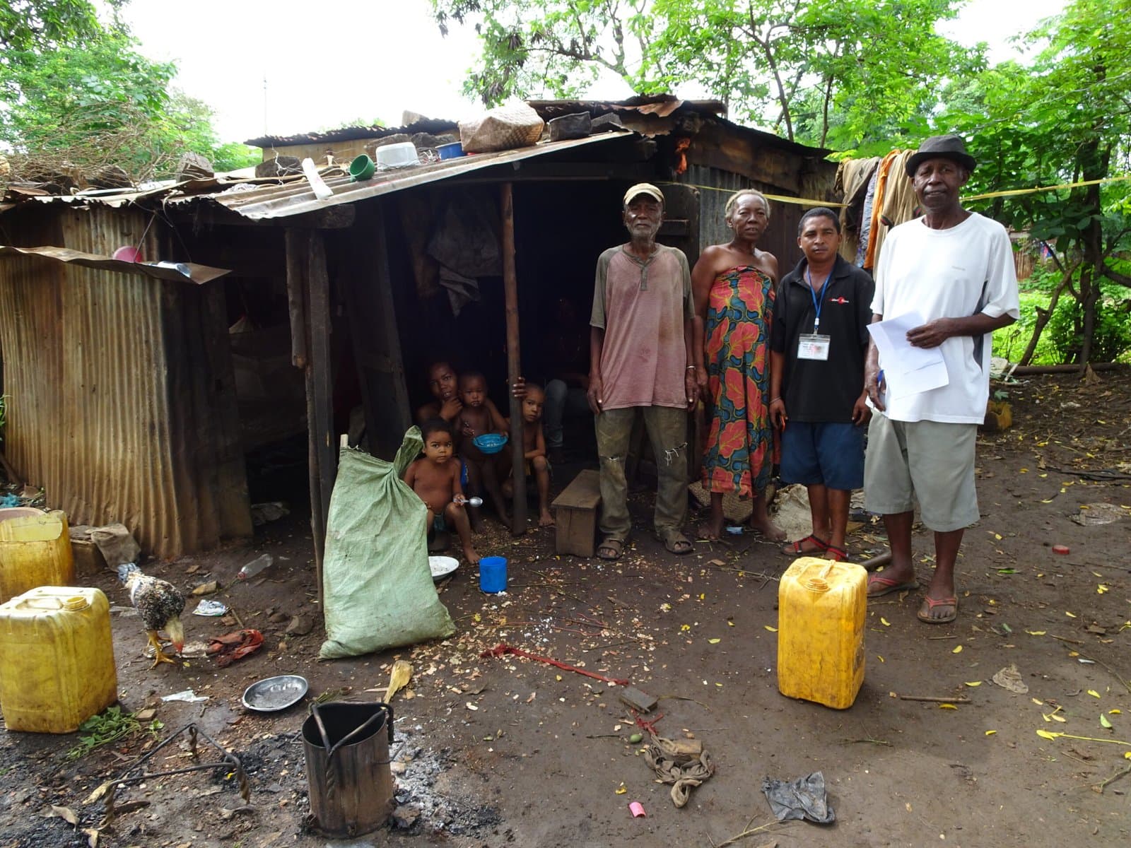 Famille debout devant sa maison en tôle et bois, entourée de bidons et d'effets personnels.