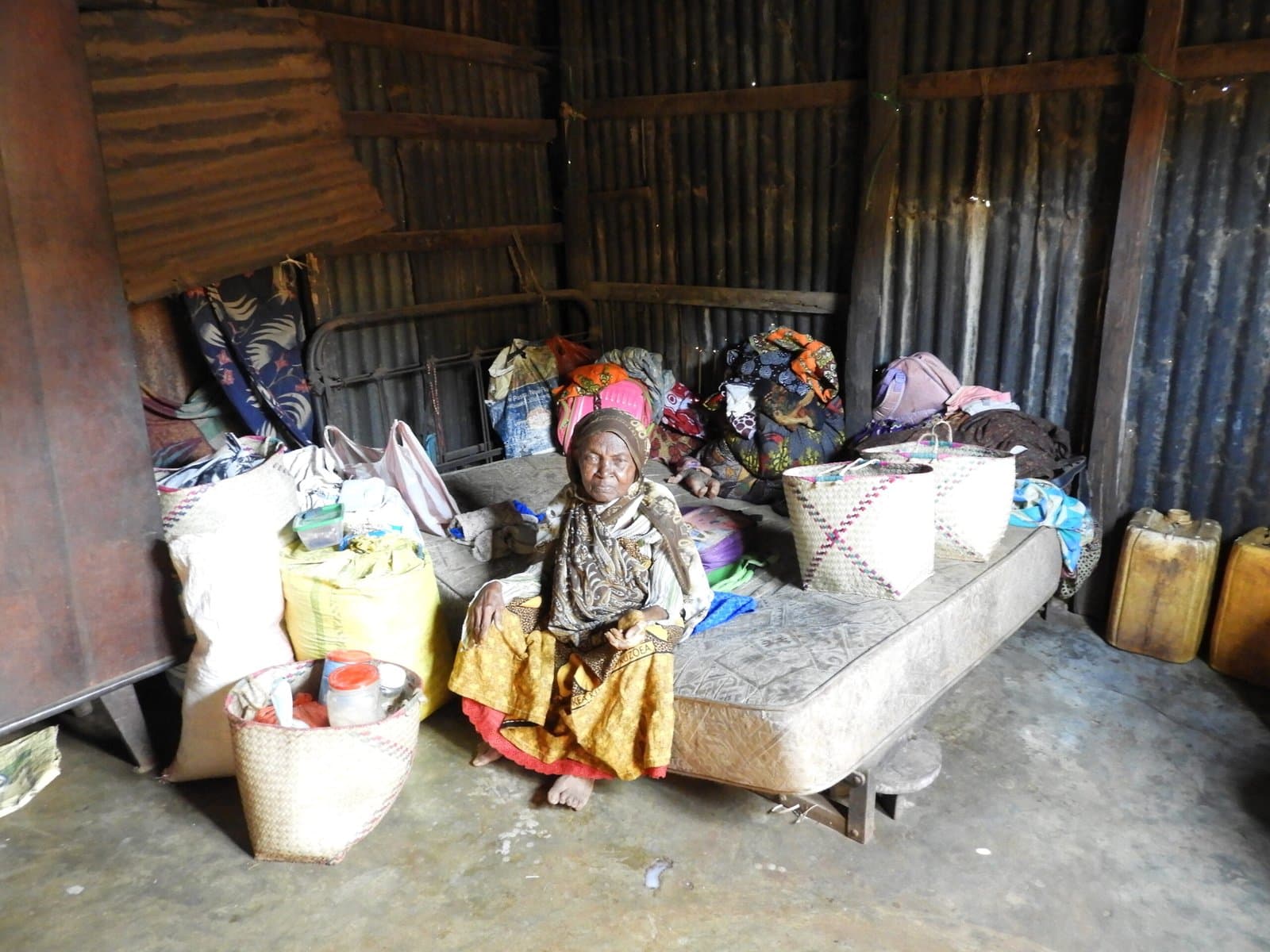 Femme âgée entourée de vêtements et d'une literie donnés, dans son habitation en tôle.