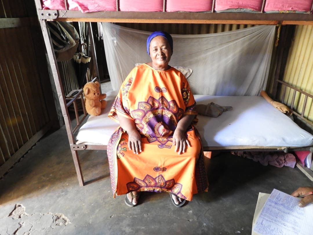 Femme âgée en robe orange assise sur son lit sous une moustiquaire.