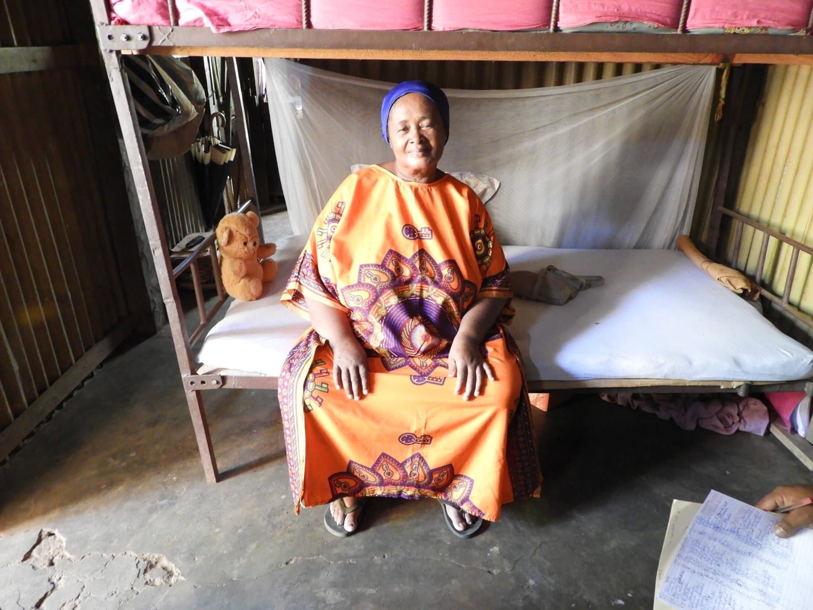 Femme âgée en robe orange assise sur son lit sous une moustiquaire.