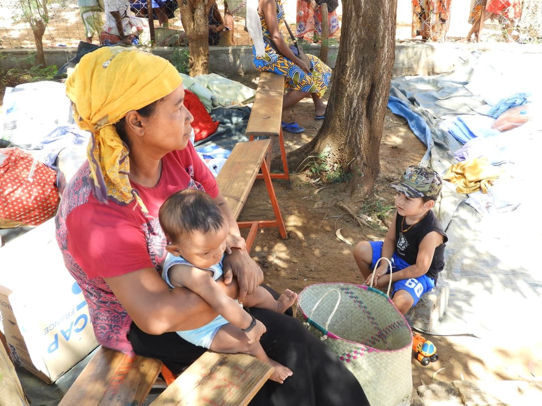 Femme portant un bébé, assise en extérieur avec un autre enfant à ses côtés.