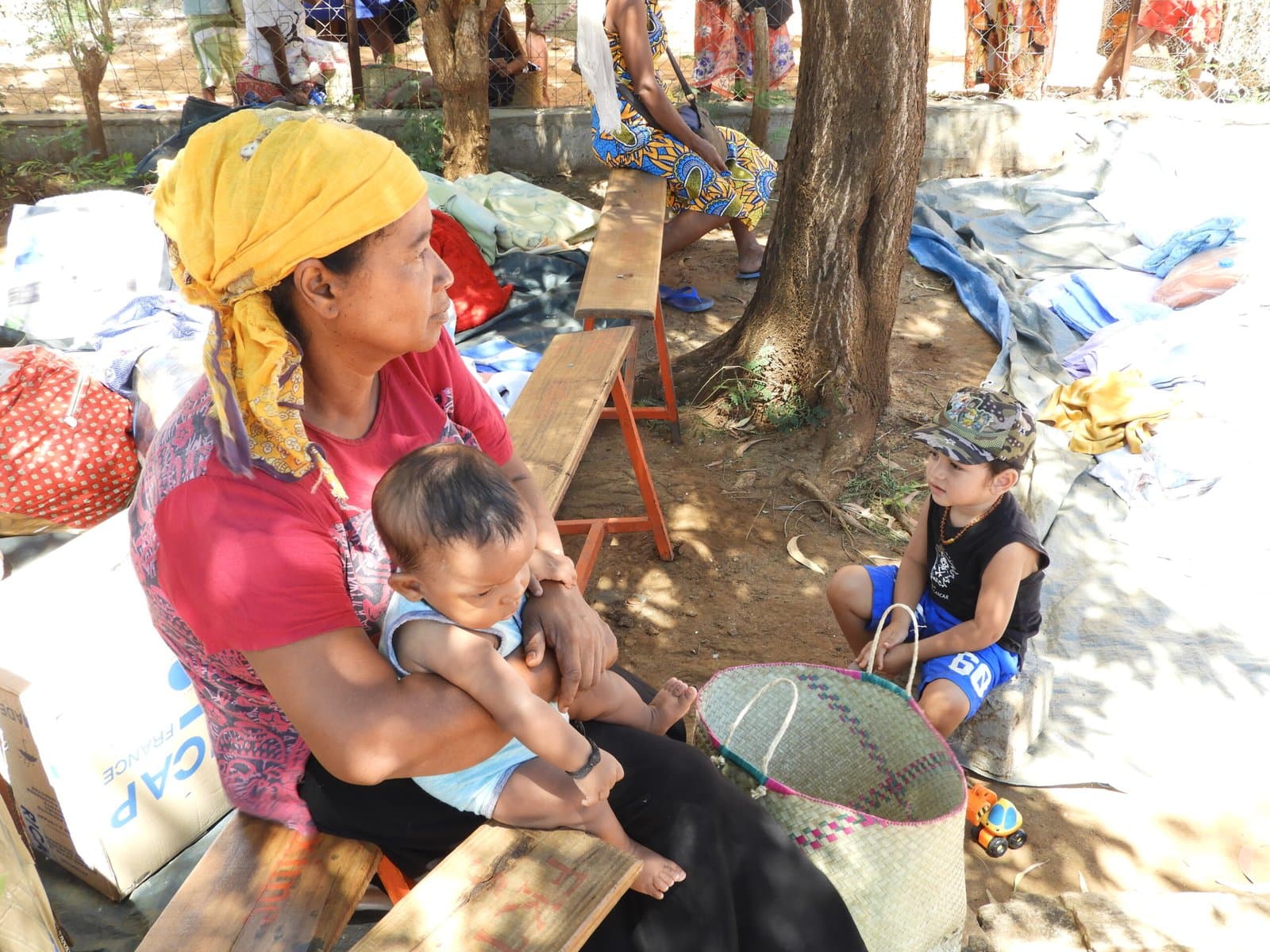 Femme portant un bébé, assise en extérieur avec un autre enfant à ses côtés.