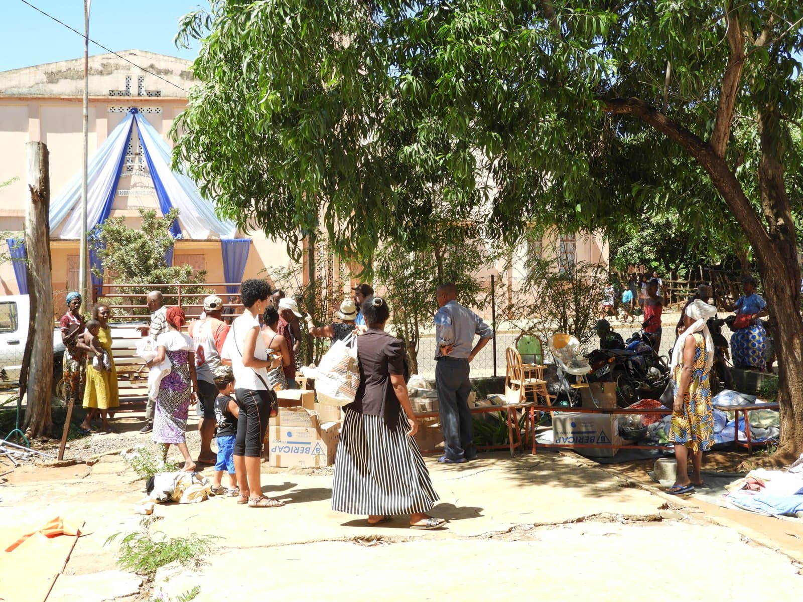 Scène de distribution sous les arbres, habitants rassemblés autour des tables d'objets donnés.