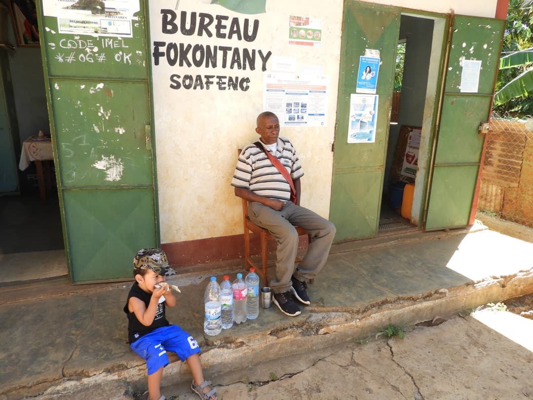 Homme assis devant le bureau du Fokontany Soafeno, bouteilles d'eau à ses pieds, un enfant à proximité.