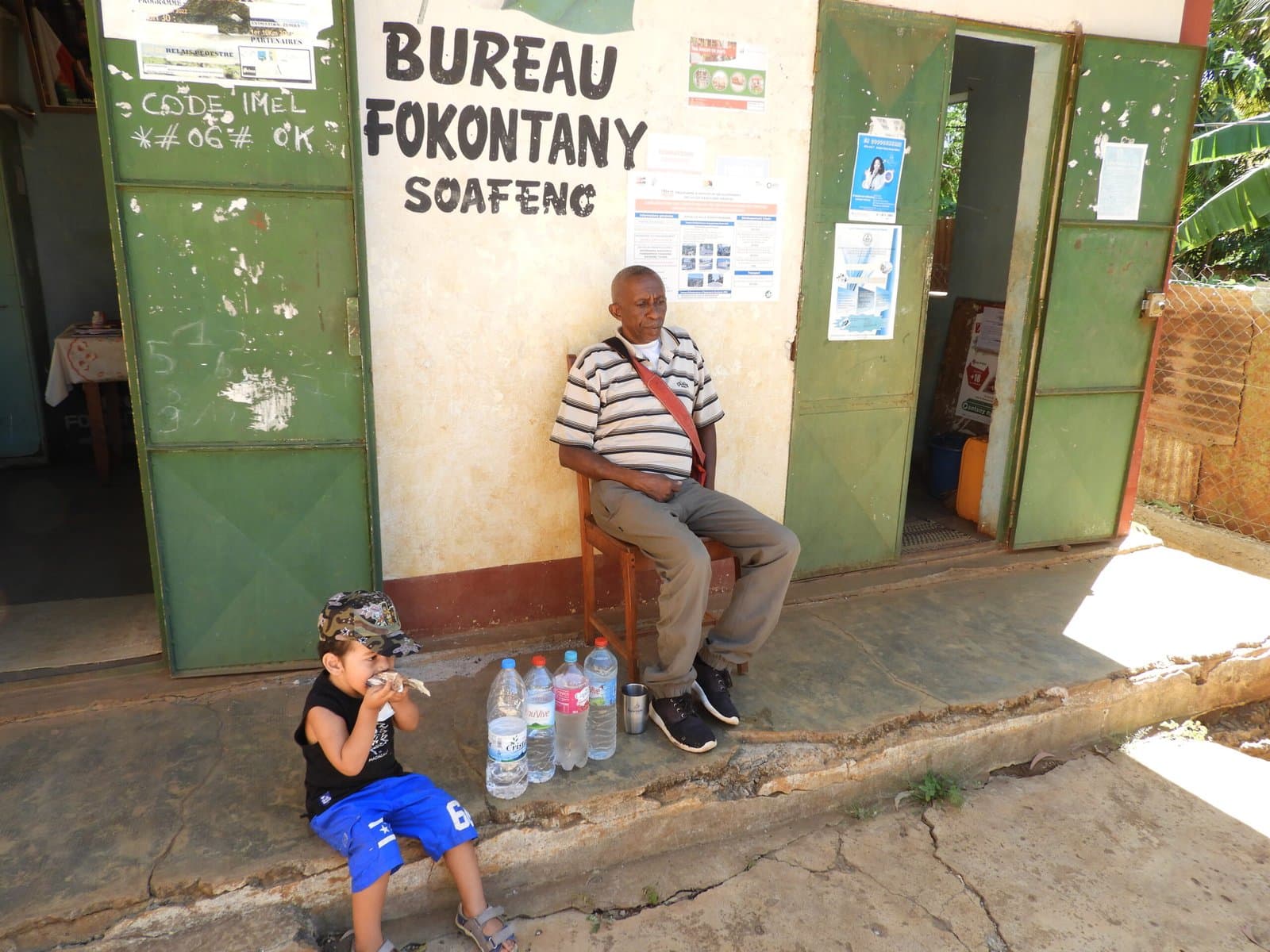 Homme assis devant le bureau du Fokontany Soafeno, bouteilles d'eau à ses pieds, un enfant à proximité.