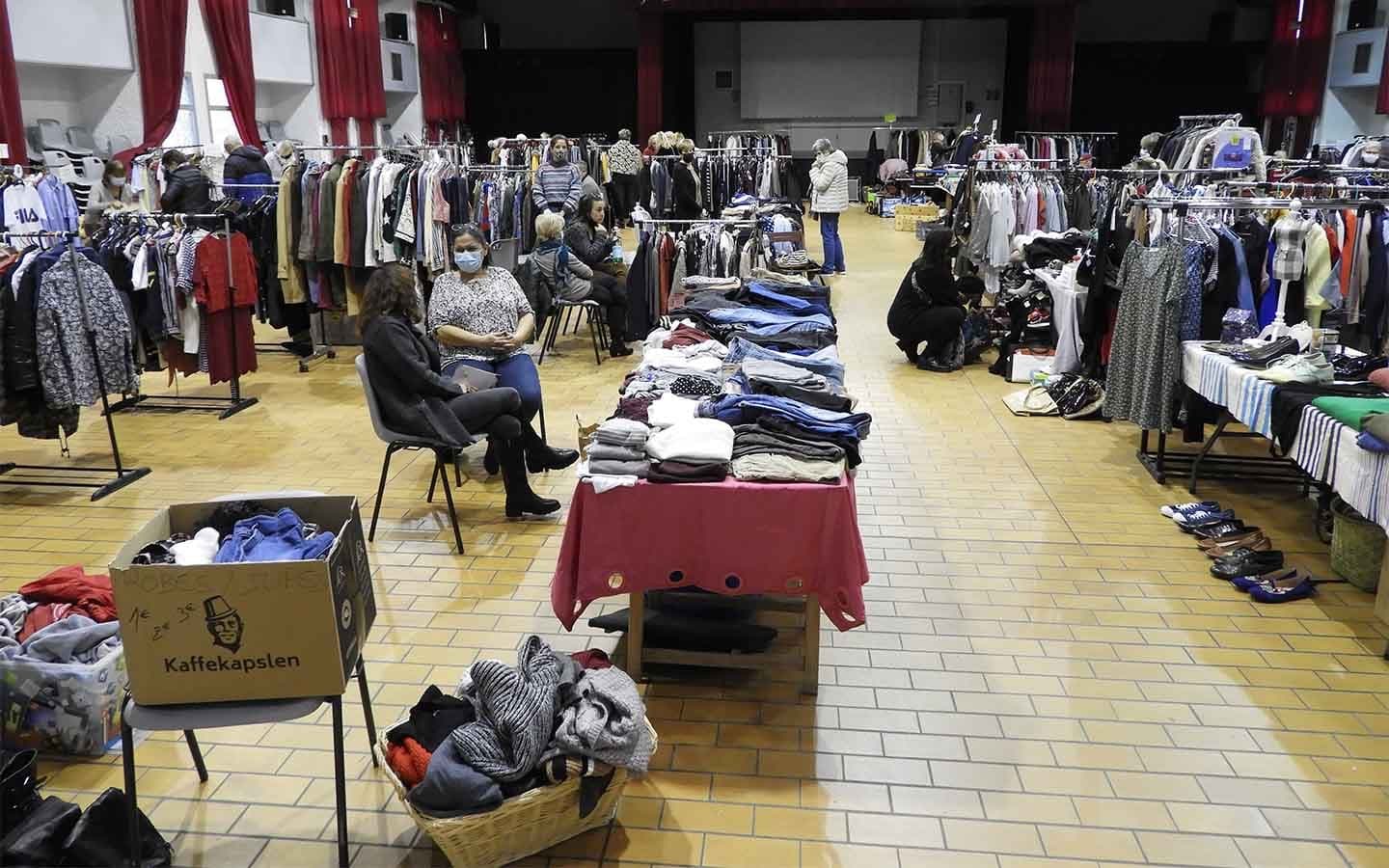 Salle des fêtes aménagée en vide-dressing solidaire, portants de vêtements et bénévoles.